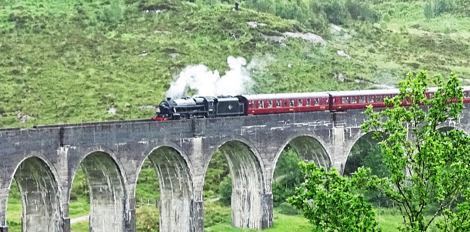 steam engine pulling box cars down a wooded track on a bridge with stone pillars