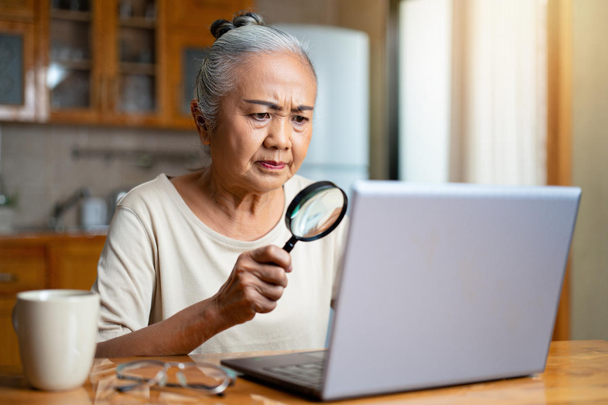 An elderly woman with a small frown squints through a handheld magnifying glass to view a laptop screen